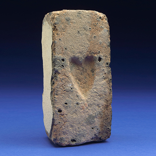 A single hand-made vintage brick of clay is stood on its end, set up under adorning studio lighting. The brick is embossed with the shape of a heart. It is a brick most likely made in Sydney, Australia, by convict labour sometime before 1840. This particular brick was offered to the market by Bonham's in New York, and sold in 2012 for US$562.50 including premium.