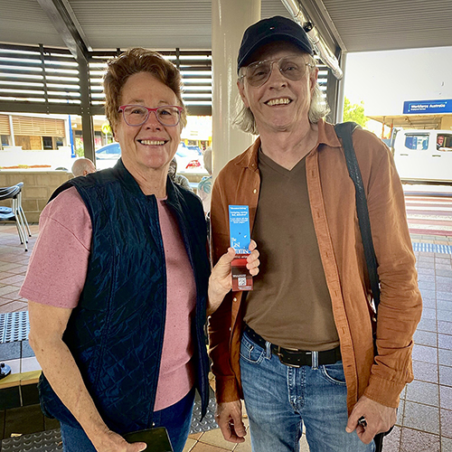 Robbie and Terrence pose for a photo, standing on the covered footpath outside the Coffee Club in Dalby. Robbie is holding up 'The Last Promise' bookmark. Smiles all round.